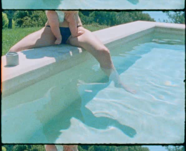 A woman in a swimsuit hugs a baby while sitting with one foot in a swimming pool outdoors during the day