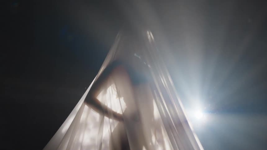 Woman stands shrouded in sheer fabric with light shining through cloth to outline her figure
