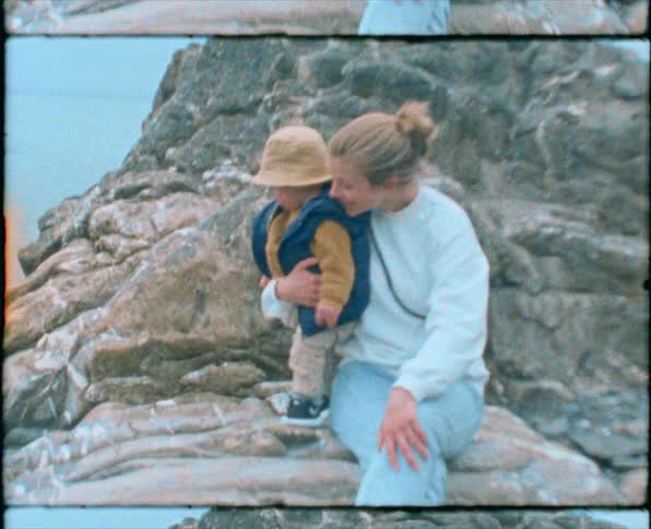 A woman holds onto her son as they sit and stand on rocks along the ocean