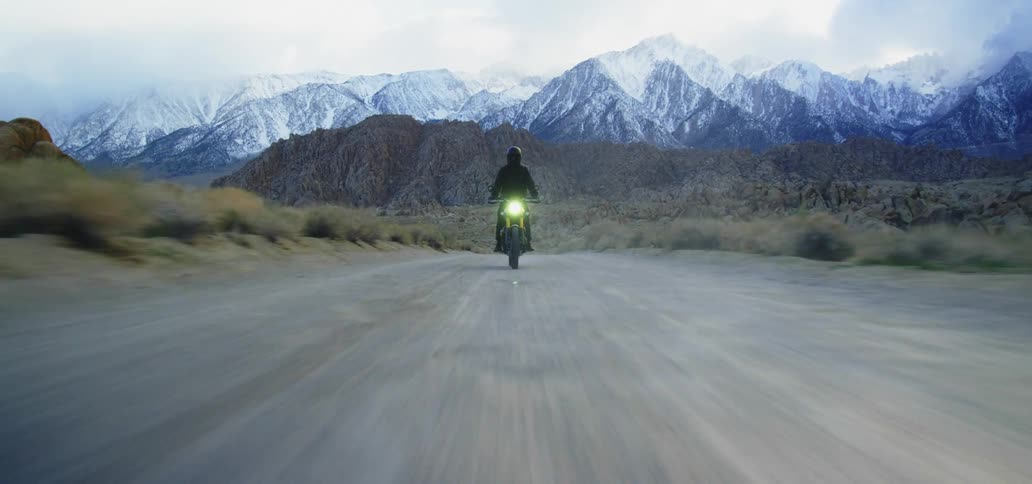 A motorcyclist rides along a road through a shrubland in the countryside while snow capped mountains loom in the distance behind them