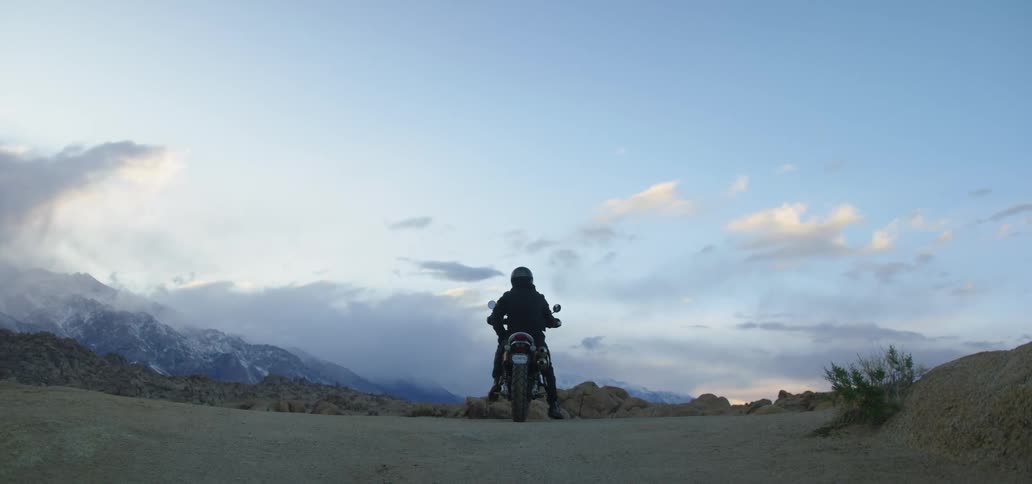Motorcycle rider sits on a motorcycle on a dirt area near the mountains