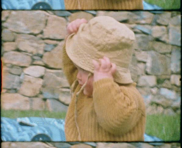 A baby boy sitting on grass and taking off a hat with a wall in the background