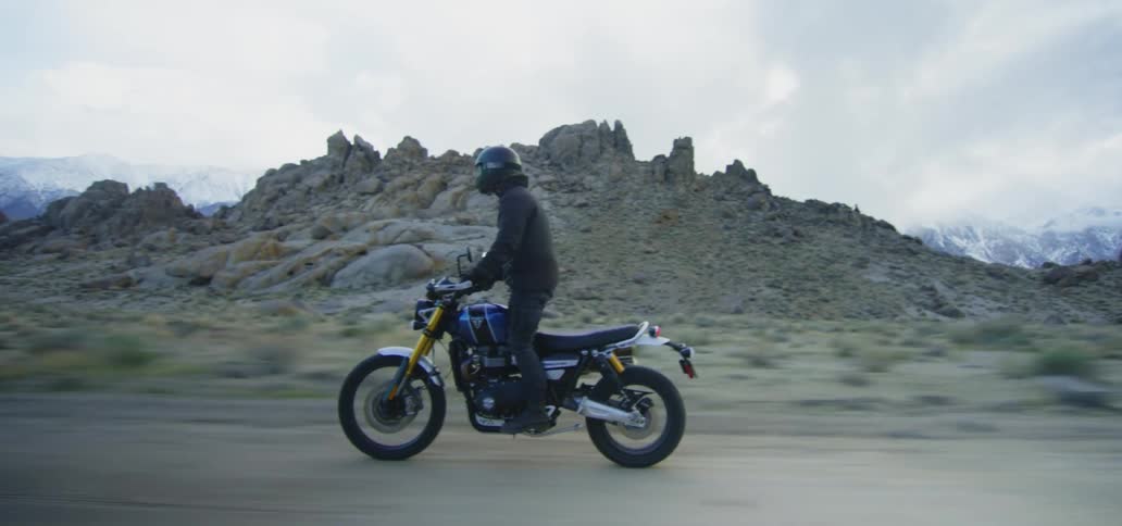 Cyclist wearing helmet stands while operating motorcycle down dirt road past craggy hills with snow covered mountains on horizon under cloudy sky