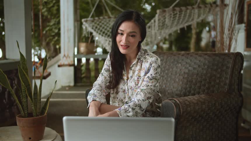 Woman with dark hair and a patterned shirt sits in brown armchair and makes a video call on grey laptop in the terrace