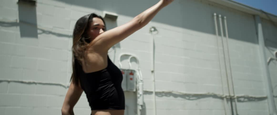 A young woman performs choreographed dance moves while outdoors next to a brick building