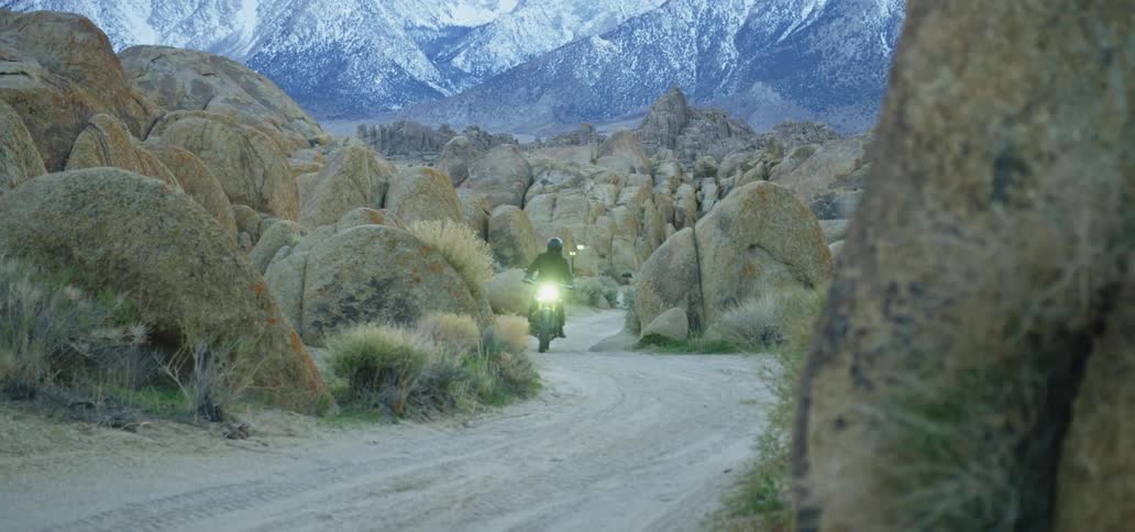 Person wearing motorcycle leathers and a crash helmet rides motorcycle along winding dirt road boulders with snow covered mountains in the background