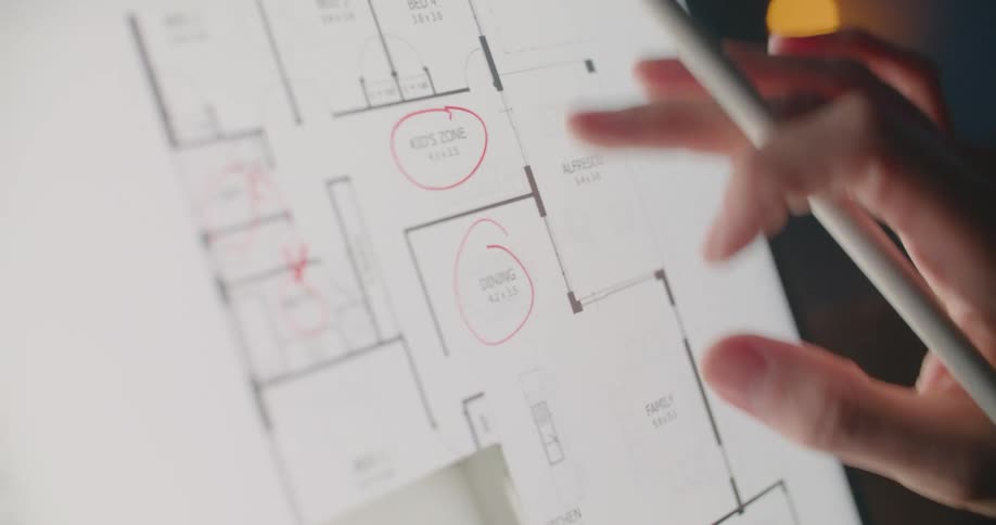 A hand holding a stylus marks a floor plan on a touch screen with a woman on video on screen