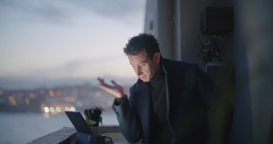 A man gestures and smiles as he talks on a tablet on a balcony