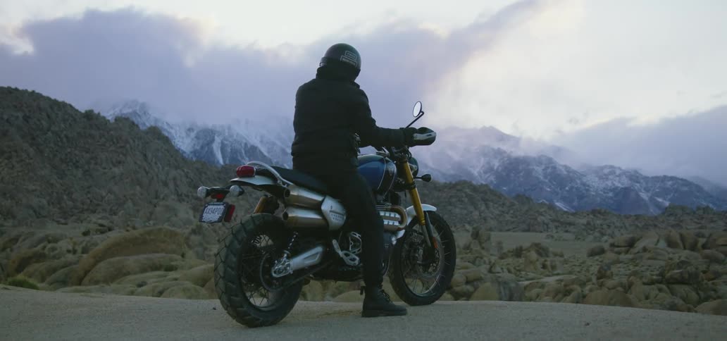 Motorcyclists sits on motorbike on edge of road looking out onto countryside bordering ice covered mountains