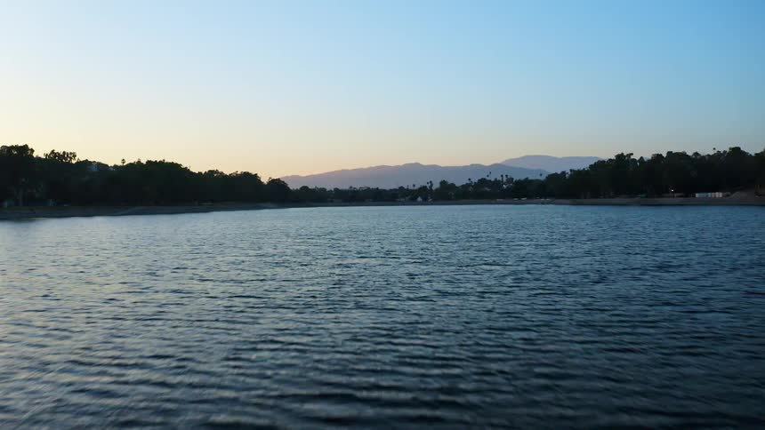 Buildings sit on shore between forest and water as mist covers mountains sitting on horizon under a red tinted sky