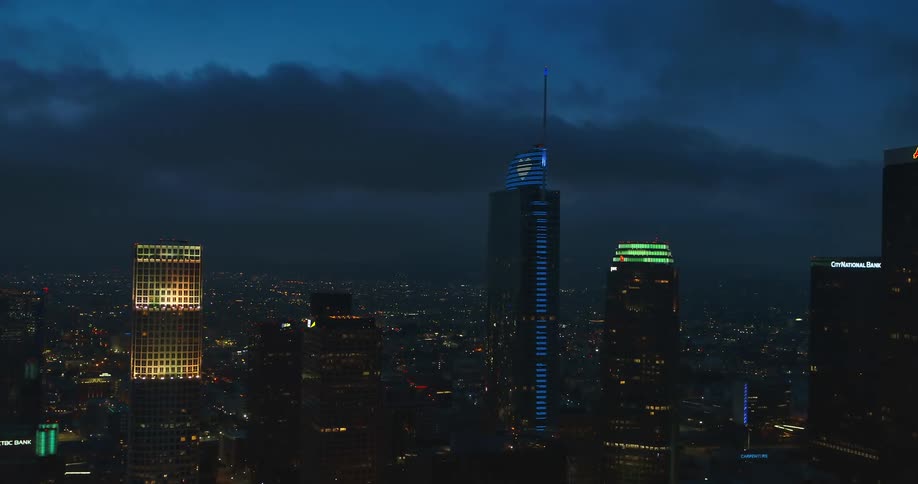 Colorful lights shine on skyscrapers in downtown high above other city lights under clouded dark blue night sky