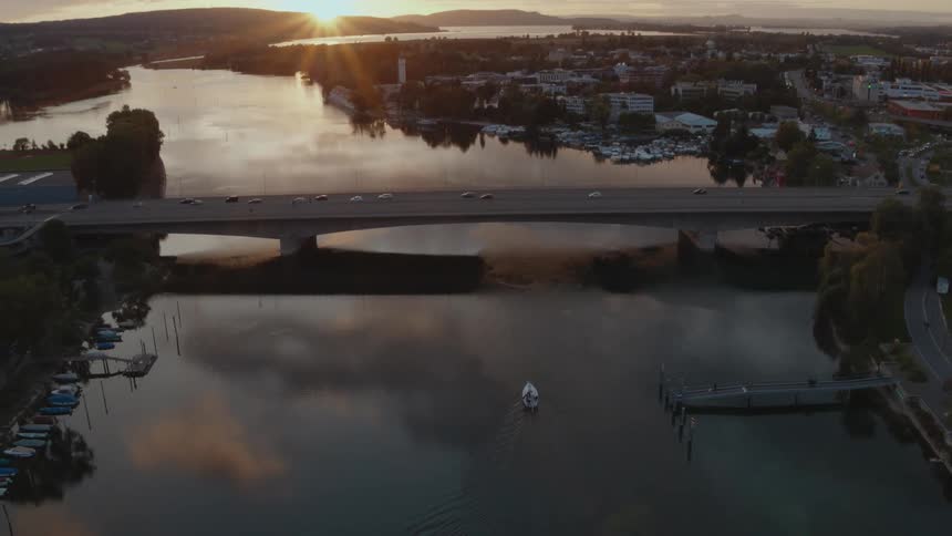 Sun peeks over mountains on horizon as lone boat move across still reflective river water toward busy bridge in middle of town