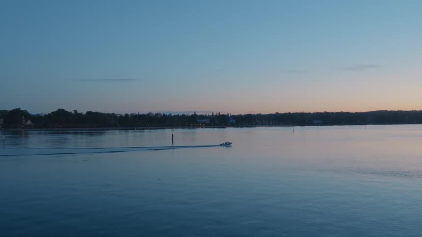 Opposing docks bookend entrance to lake as motorboat parallels forest covered shore leaving a wake on rippling reflective water under a red tinted sky