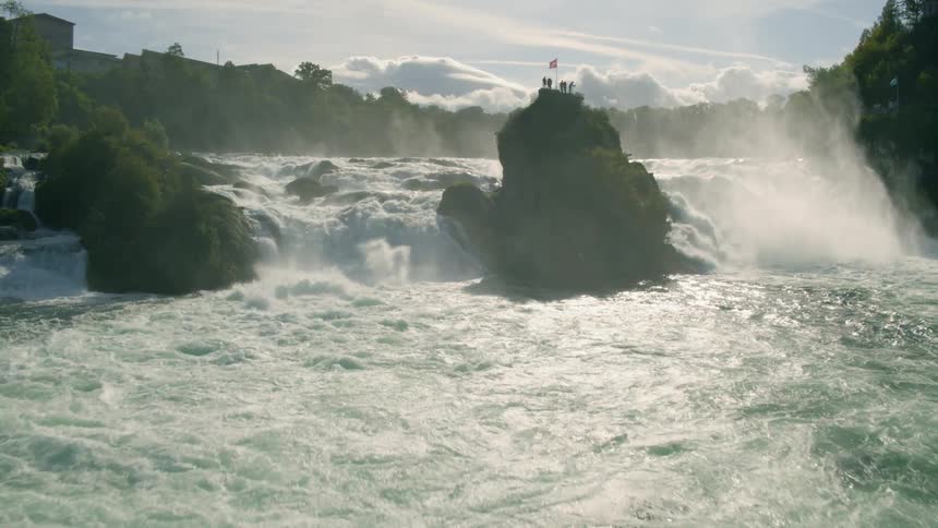 A river with a boat a waterfall and a bridge with people standing on a platform with the sky and clouds in the background