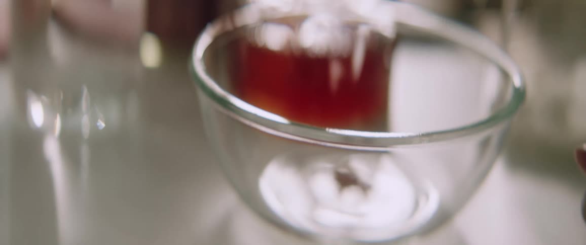 A woman adds a tablespoon of brown powder to a glass mixing bowl in a lab