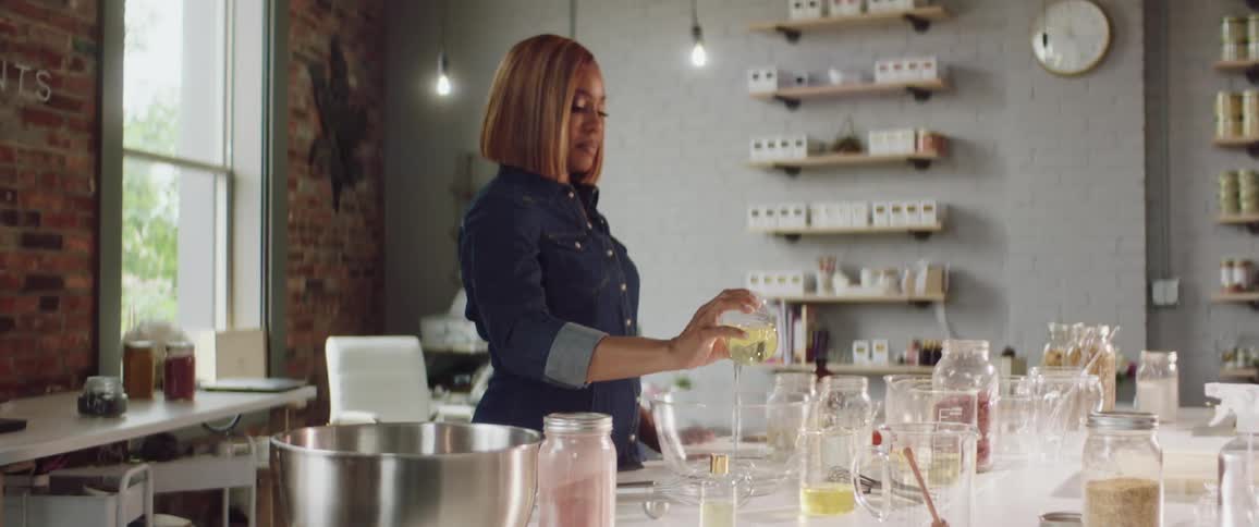 A woman pours the contents of a beaker into a large glass bowl while mixing oils
