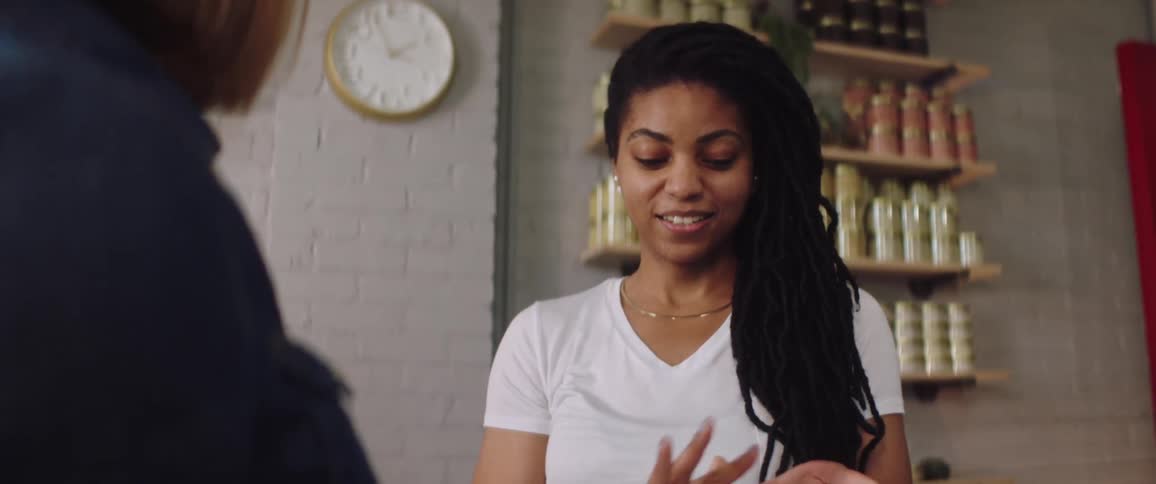 Two women in a room with one smelling and rubbing cream on her hands with a wall shelves and a clock in the background