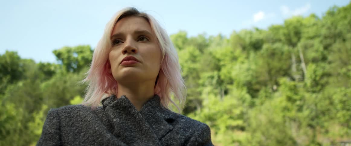 Blonde woman with tinted hair walks near woods under blue sky