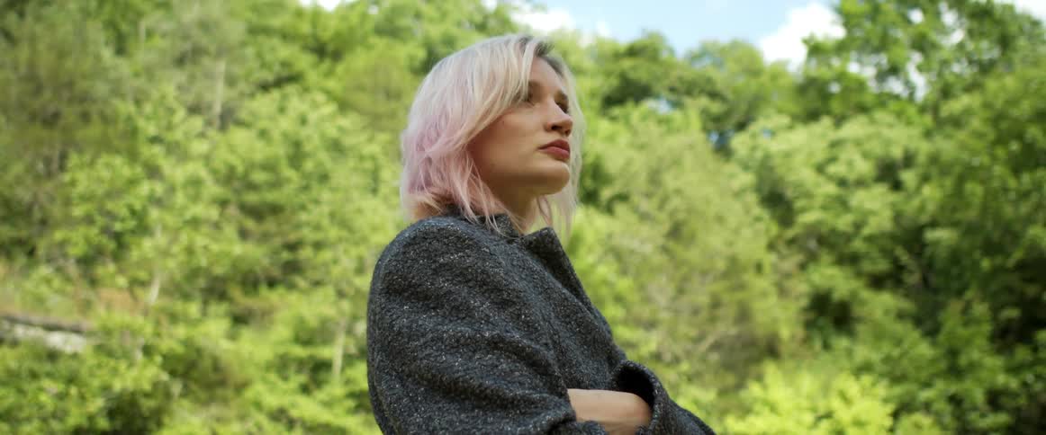 A woman with blonde hair stands outdoors in a clearing near a forest