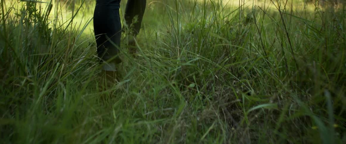 A person in boots walks through tall grass in a field during the day