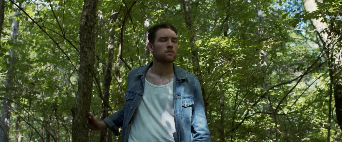 Bearded man in a denim jacket touches a tree trunk as he walks through the forest