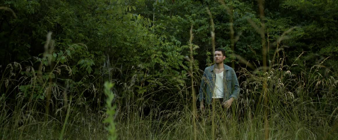 A man with a denim jacket walking on a landscape with grass with trees in the background
