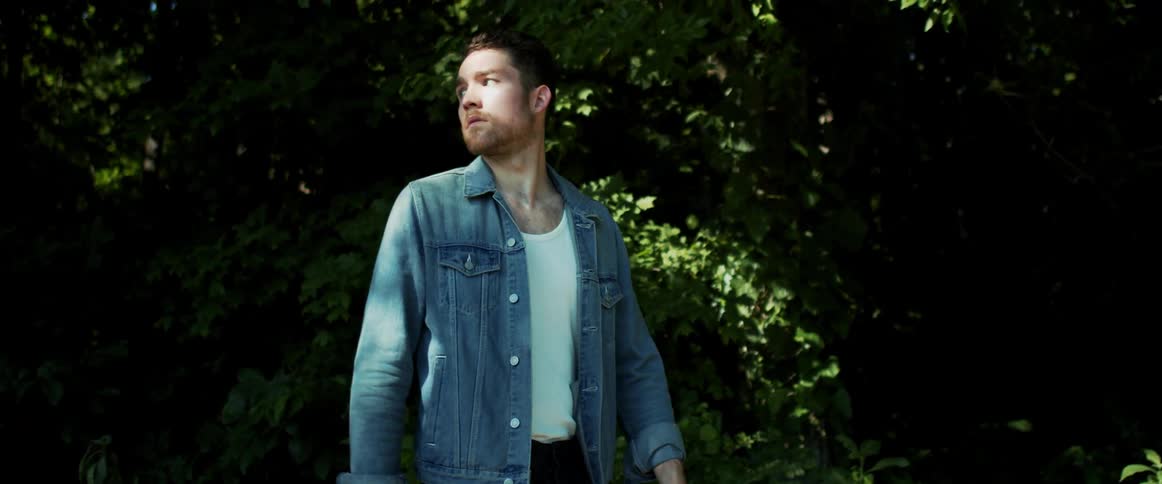 A man with a denim jacket walking with trees and plants in the background