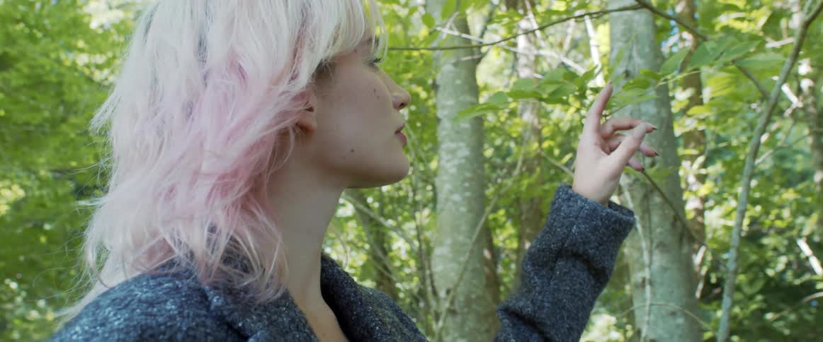 A woman touches tree branches and leaves as she walks through a forest