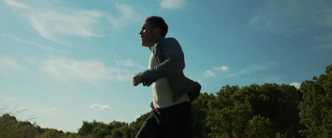 Man wearing jean jacket and tank top runs near forest under lightly clouded sky