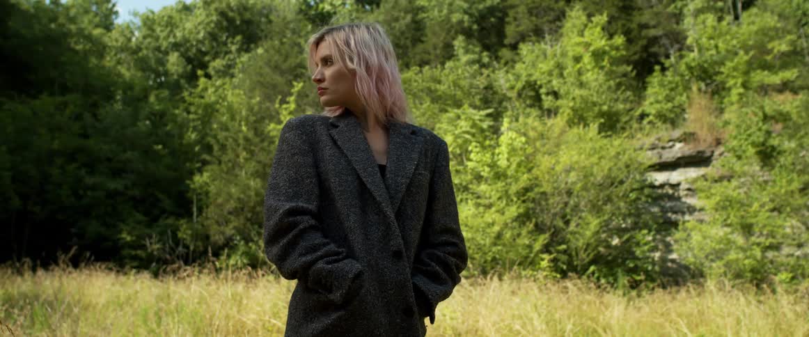 Blonde woman with pink tinted hair wearing dark gray coat walks through field near woods stopping to look back briefly