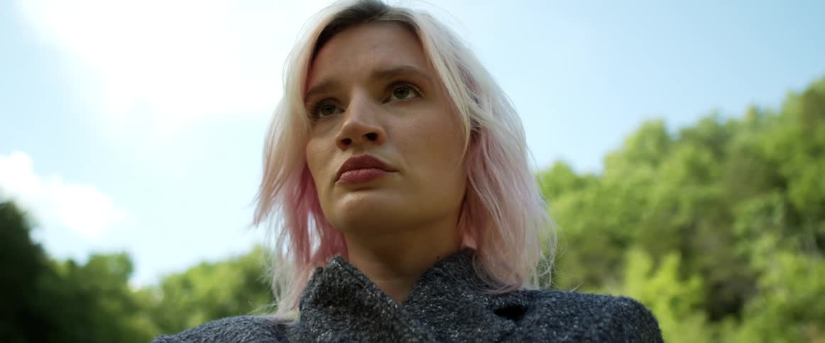 A woman with dyed blonde and pink hair stands and stares outside with trees behind her