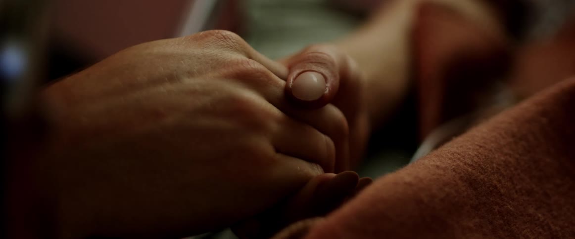 A womans hand holds and squeezes another as she lies in a bed covered by a blanket