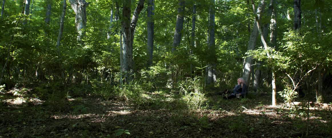 A depressed woman sits against a tree in a forest during the day