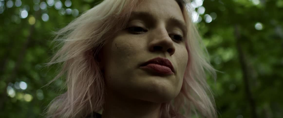 A woman standing within a grove of trees lowers her gaze as she looks pensive