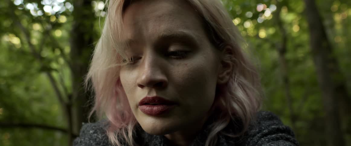 Blonde woman with magenta tinted hair looks downcast in forest