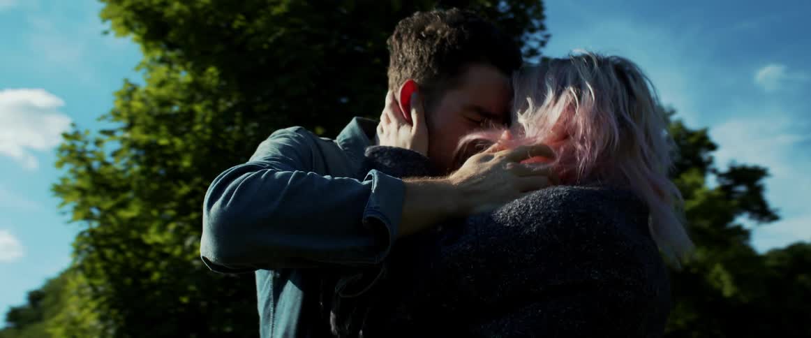 A man and woman stand touching foreheads and caressing faces outside on a bright blue sky day