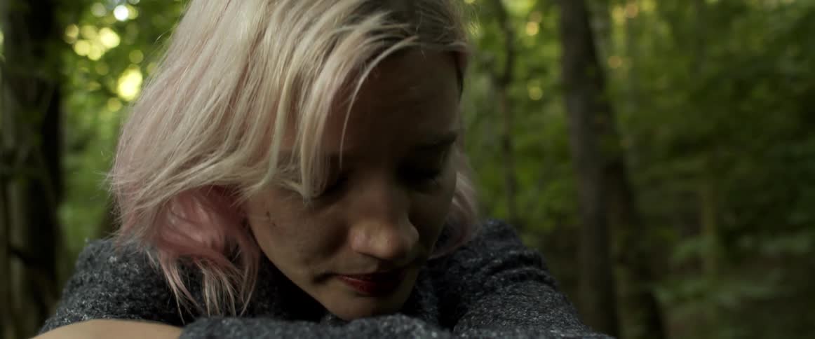 A woman sits in a forest with arms wrapped around herself looking despondent