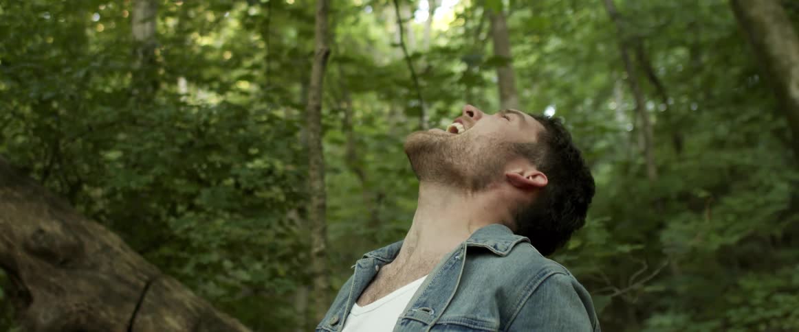 A man with a denim jacket in a forest with trees the sky and clouds in the background
