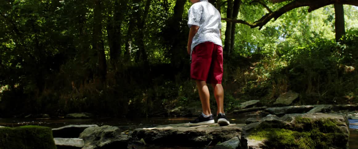 A boy jumping on rocks with a stream trees the sky and clouds in the background