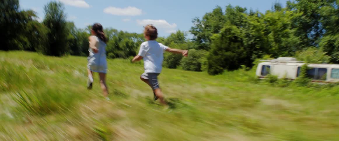 Kids play tag in field near parked rv bordering woods under slightly clouded sky