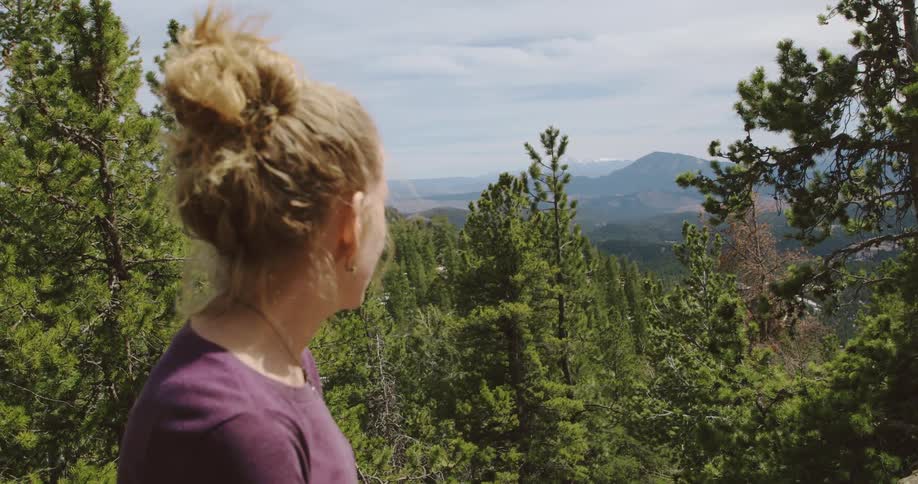 Blonde woman standing in front of a forest in the mountains at daytime