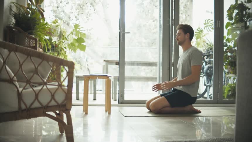 A man sits back on his heels as he kneels on a mat at home preparing to meditate