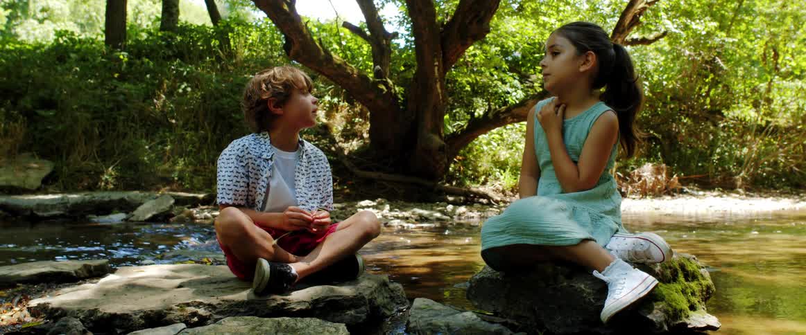 A young girl twirls her ponytail as she sits on a rock by a stream talking to a boy