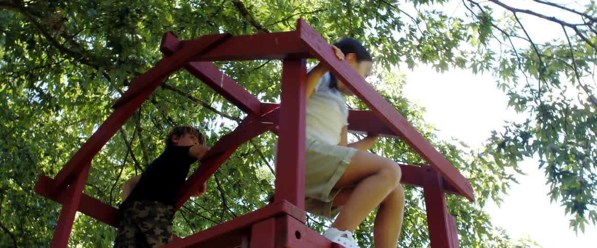 A boy and a girl playing in a jungle gym with trees the sky and clouds in the background