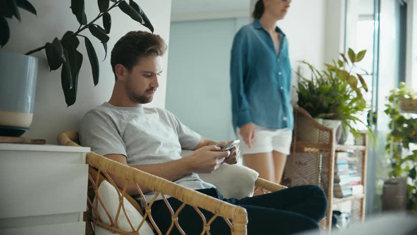 A man using a smartphone indoors at home while a woman walks by