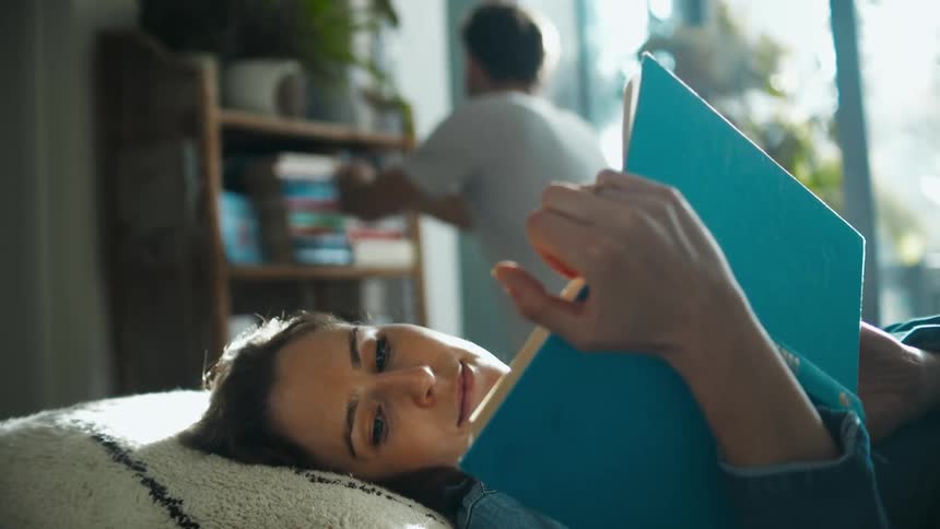 A woman reads a book while lying on a pillow as a man checks books on a bookshelf behind her