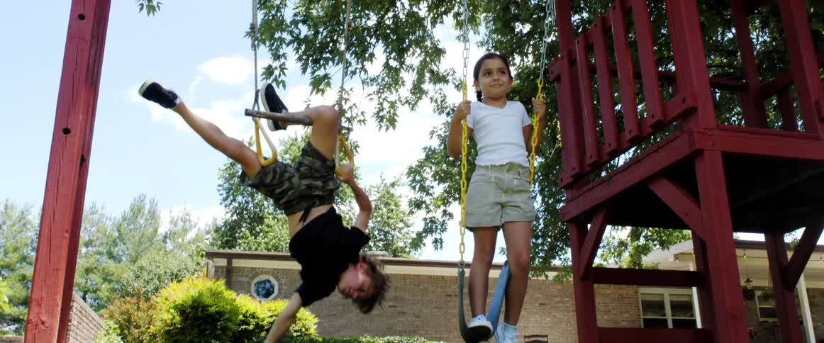 A girl stands on a swing while her brother flips upside down on another