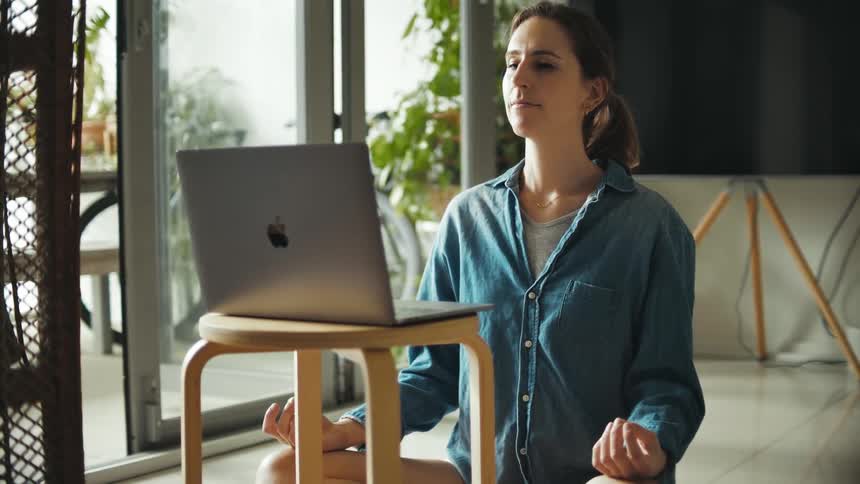 A woman practices yoga on the floor while sitting before a laptop on a stool