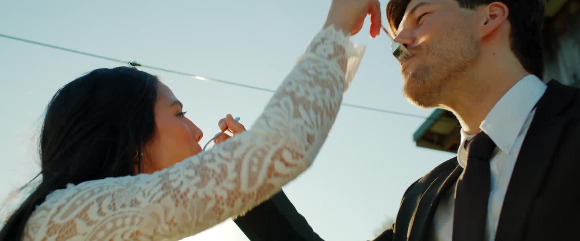 A man and woman in formal wear feed each other with forks while standing outdoors on a sunny day