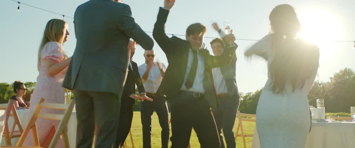 A man in a suit cheerfully dances in the center of a circle formed by cheering people in formal wear at a garden party during the day